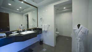 Une salle de bain moderne avec un grand miroir, un lavabo et des toilettes. Une robe est accrochée au mur et une poubelle se trouve près du lavabo.