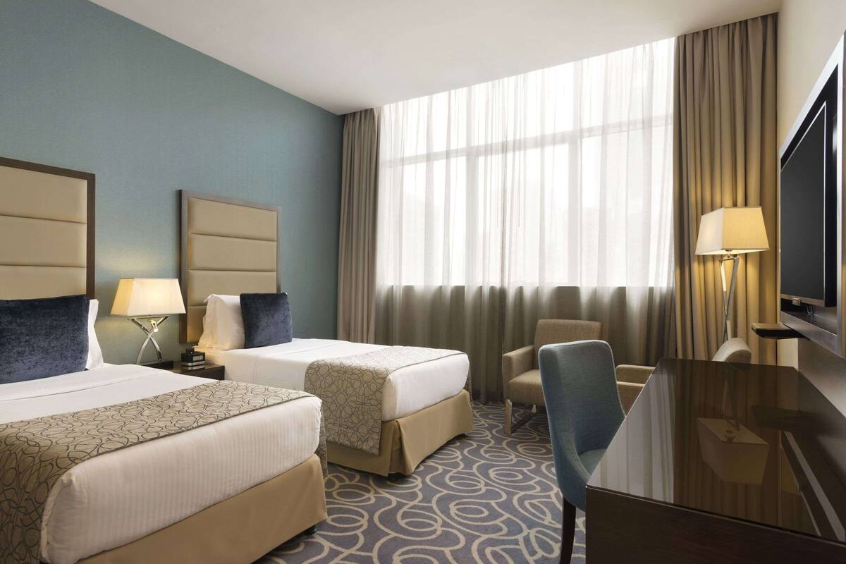 A hotel room with two beds, a desk, a chair, a TV, and a large window with curtains.