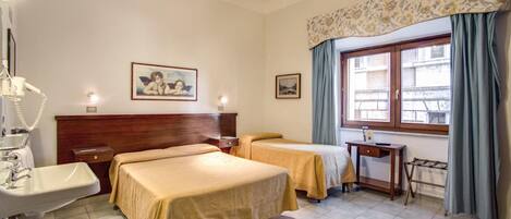 A hotel room with two beds, a bedside table, a desk, a chair, a window with curtains, and a wall-mounted lamp.