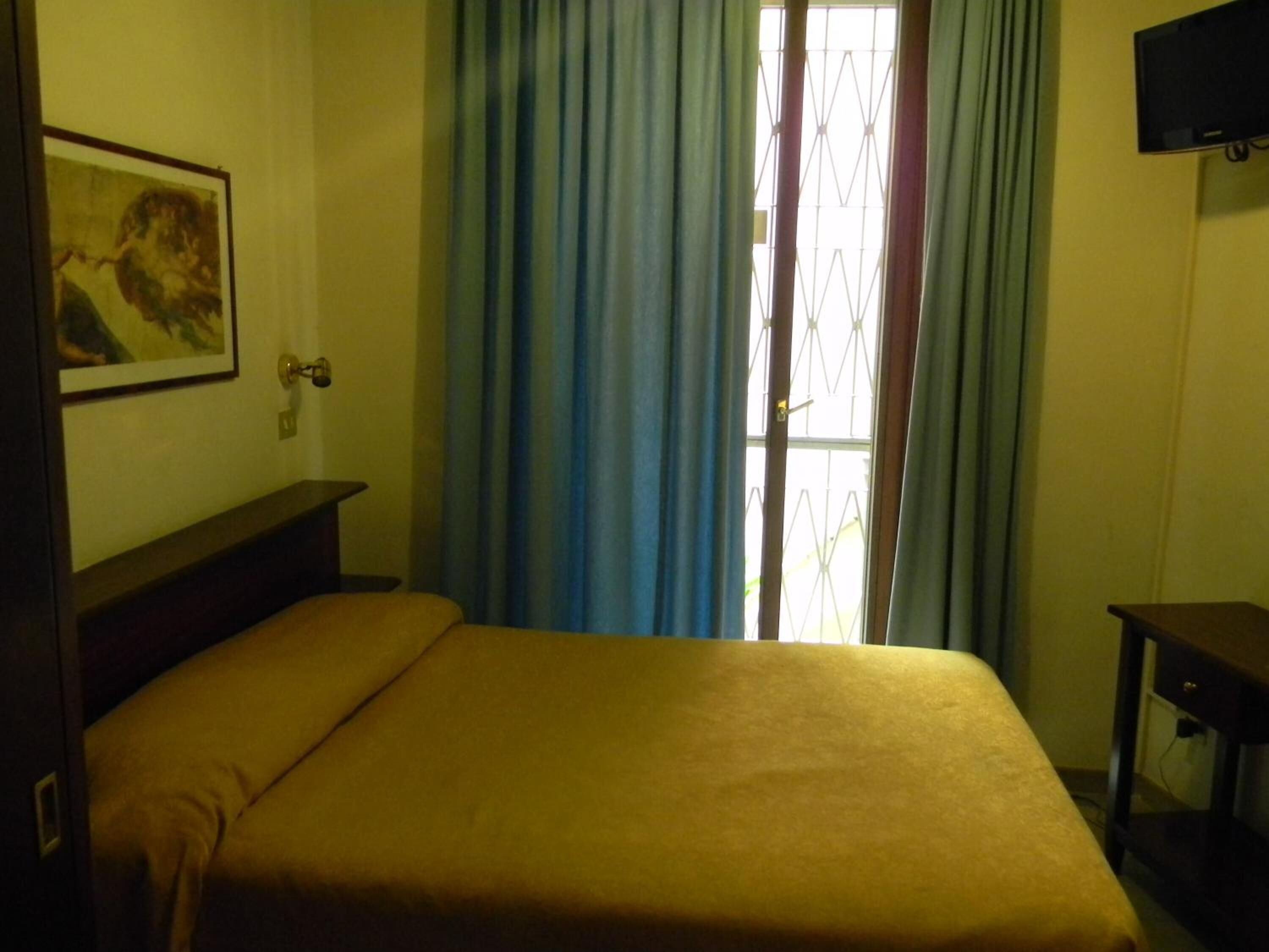 Une chambre avec un lit, une petite table, un téléviseur fixé au mur et une fenêtre avec des rideaux.