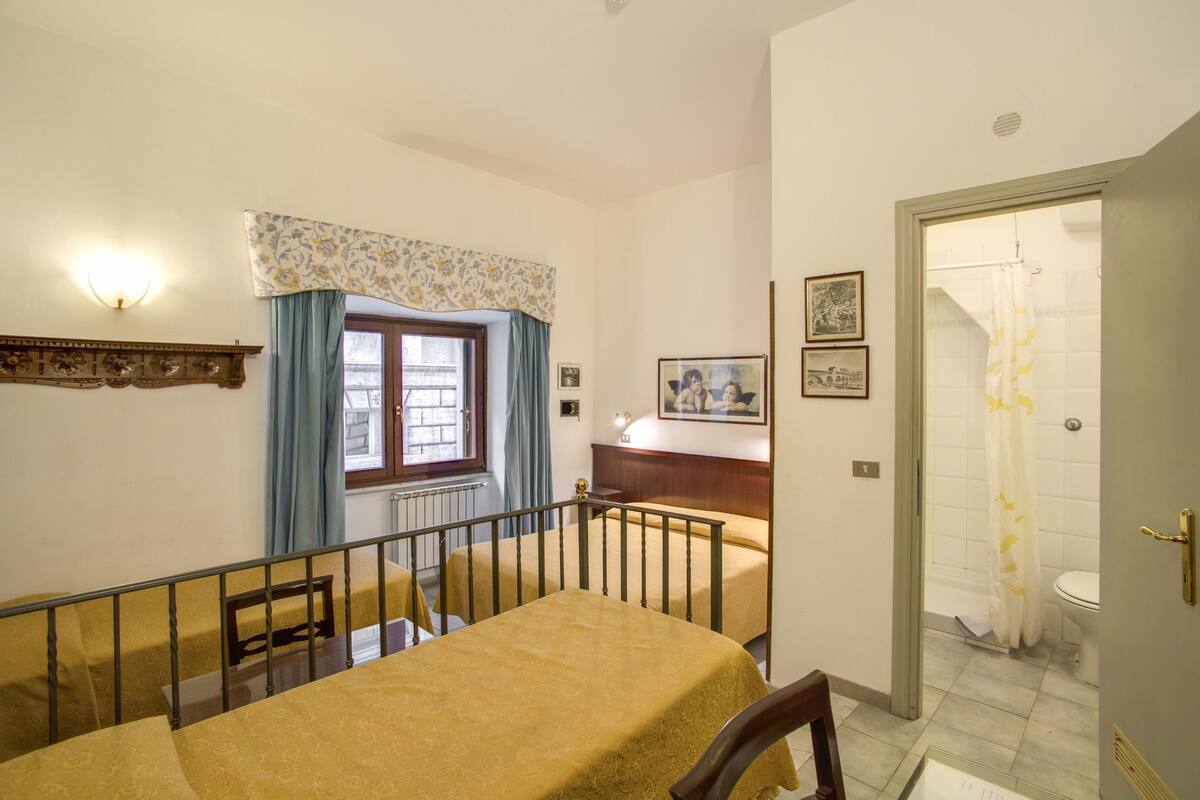 A hotel room with a bed, a window with curtains, a shower area, and a small table with a chair.