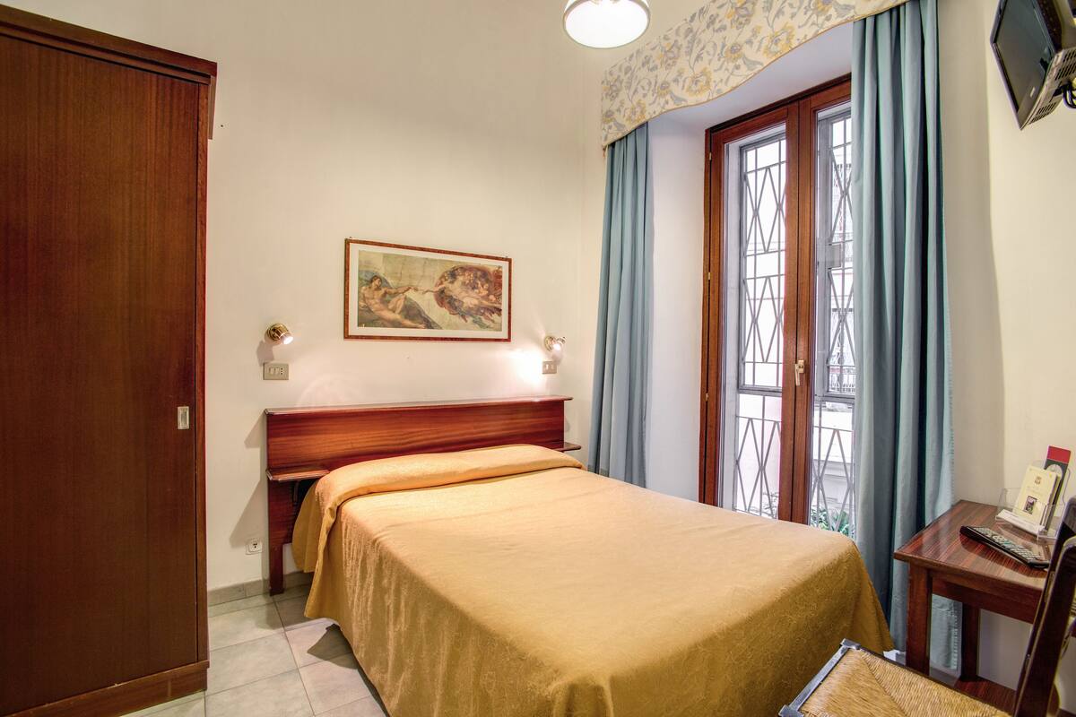 A hotel room with a bed, a wooden wardrobe, a desk with a chair, a window with curtains, and a painting on the wall.