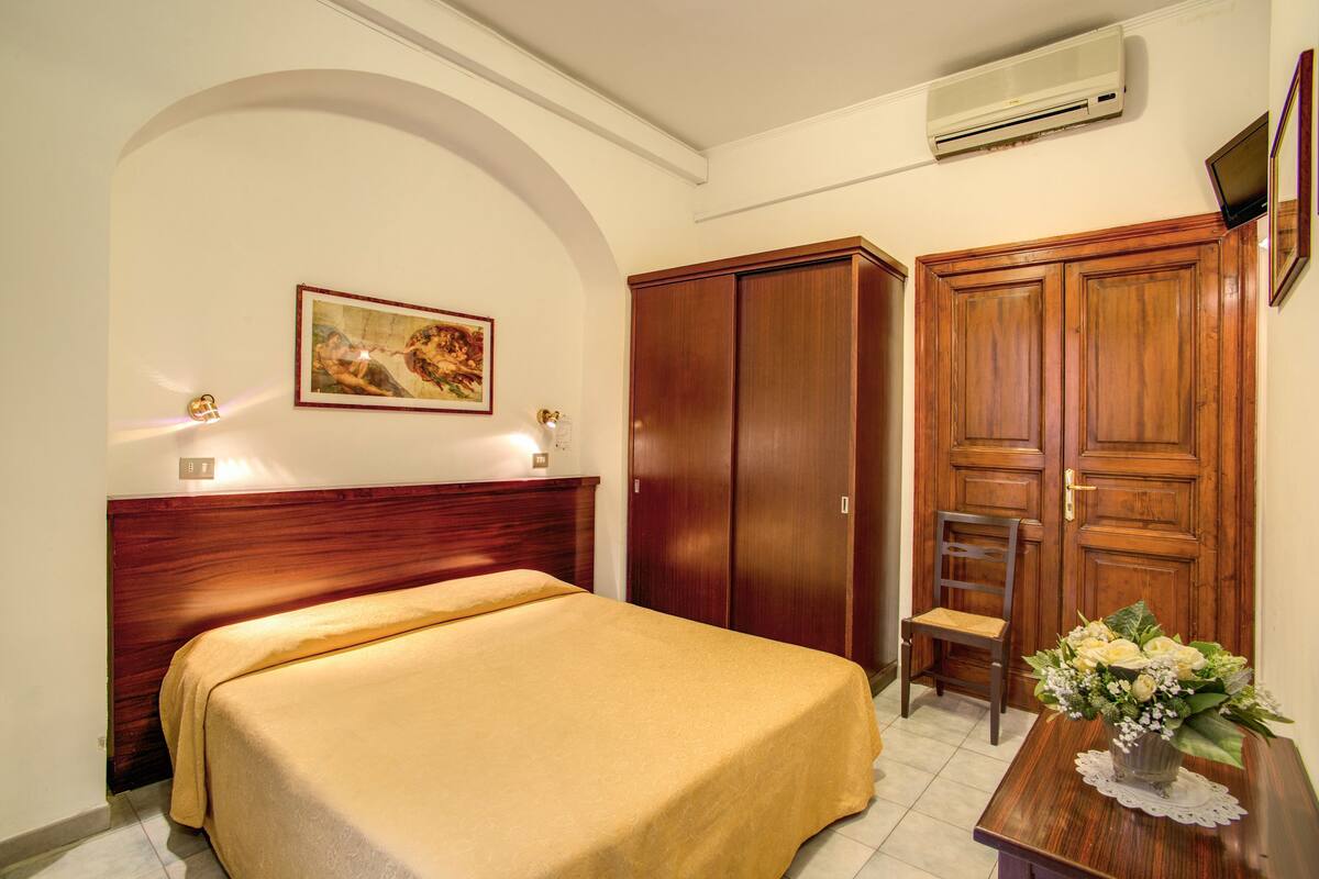 A hotel room with a bed, a wooden wardrobe, a desk with a vase of flowers, and a painting on the wall.