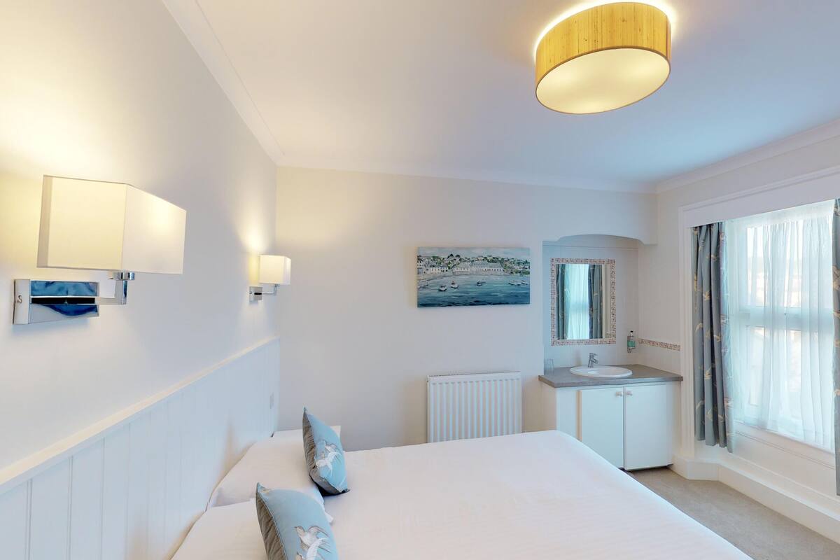 A hotel room with a large bed, a bedside table, a mirror, a window with curtains, and a painting on the wall.