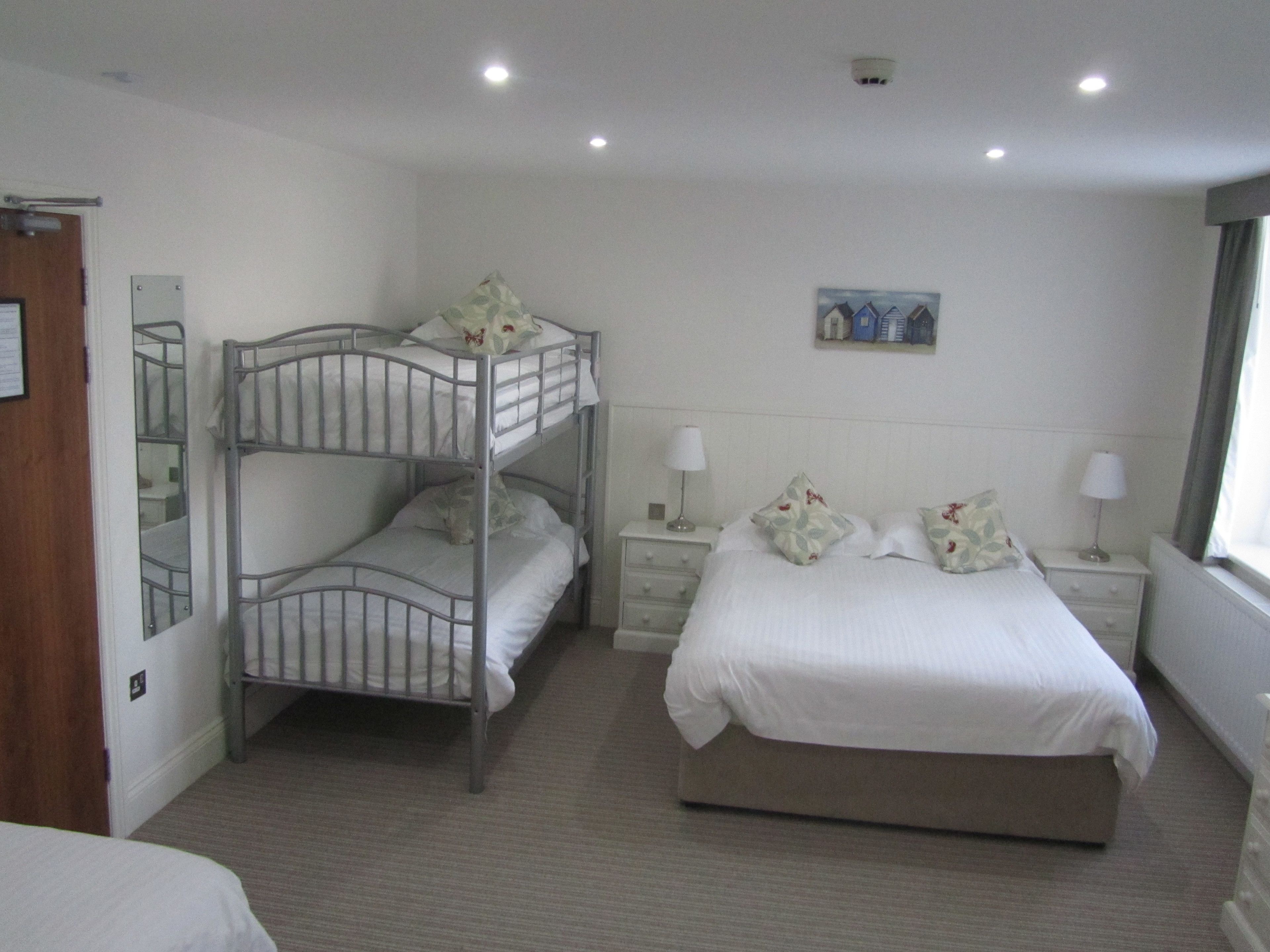 A hotel room with bunk beds, a single bed, a nightstand with lamps, a painting on the wall, and a window with curtains.