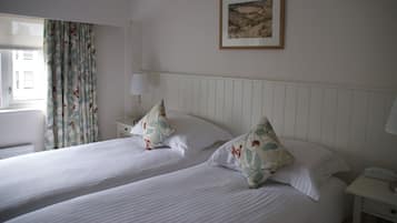 A hotel room with two beds, a window with curtains, a bedside table with a lamp, and a framed picture on the wall.
