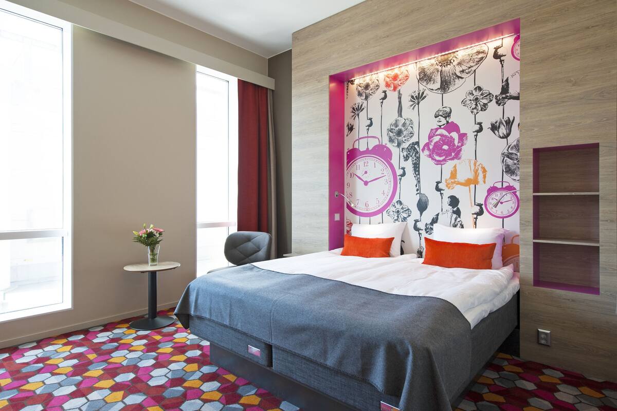 A modern hotel room with a large bed, a small side table with a vase, a chair, a wooden headboard with floral wallpaper, and a colorful hexagonal carpet.