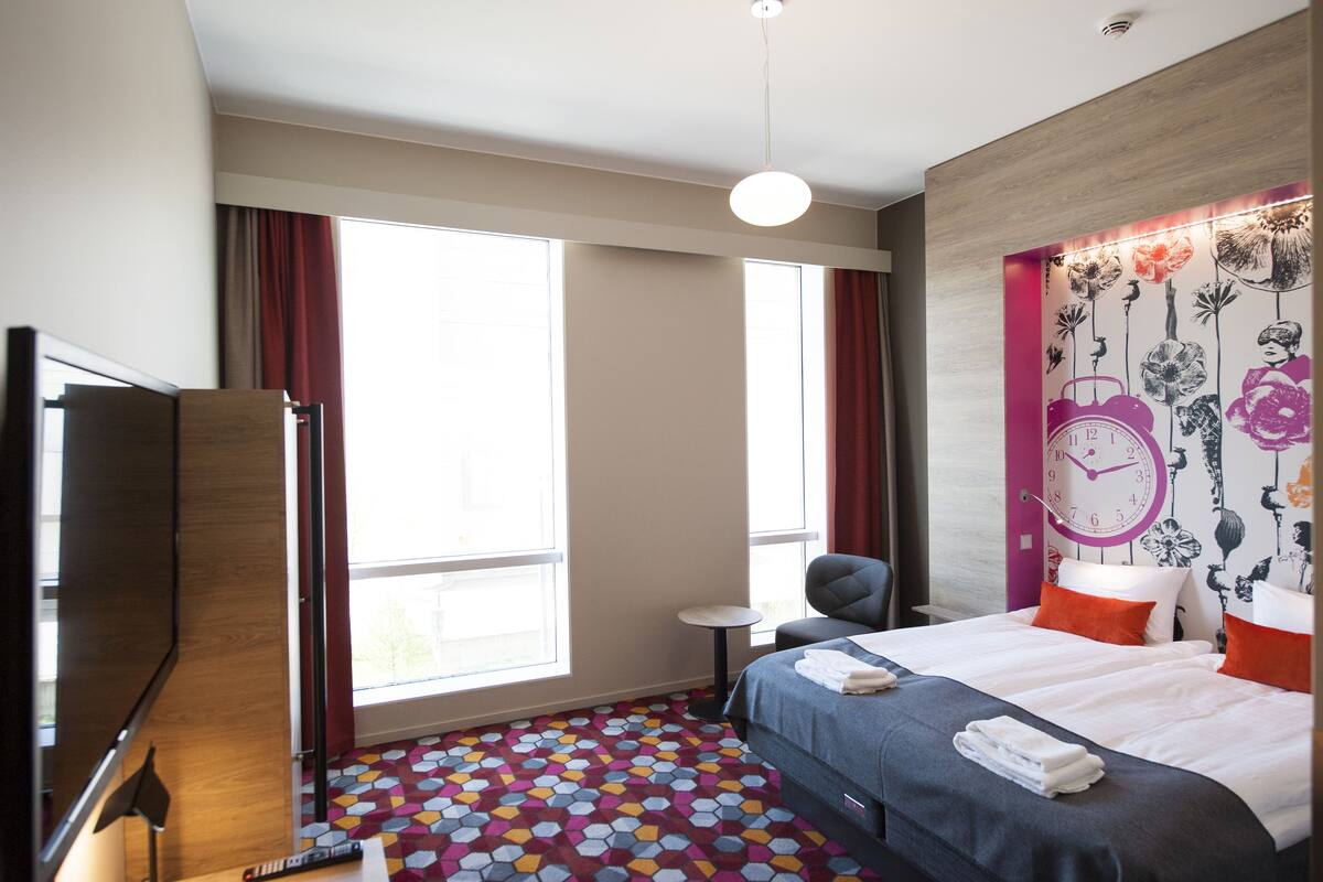 A hotel room with a large bed, a small desk, a chair, a TV, and a window with curtains.