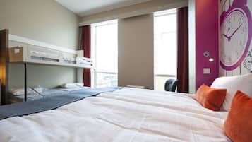 Hotelkamer met een stapelbed, een groot bed met wit beddengoed, oranje kussens, een wandklok en een raam met gordijnen.