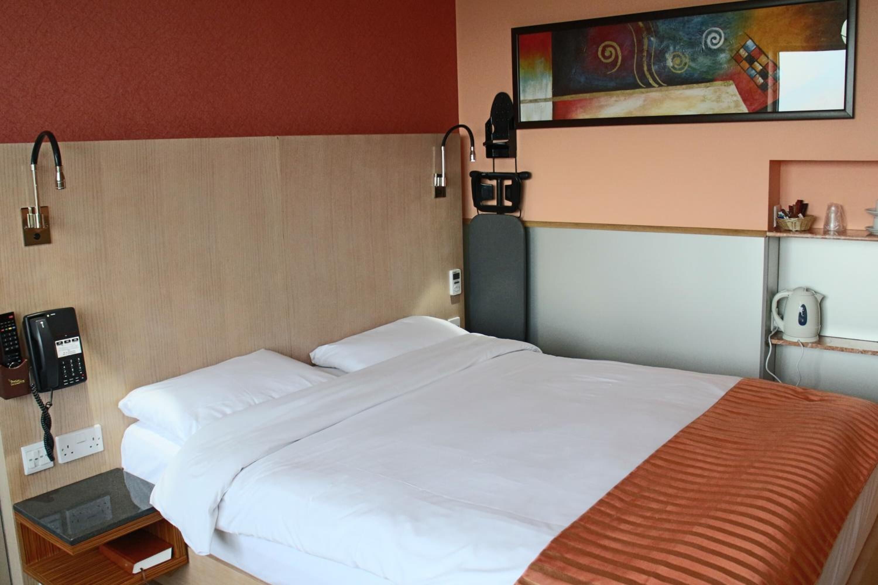 Habitación de hotel con cama, escritorio con teléfono, estante con hervidor y un cuadro en la pared.