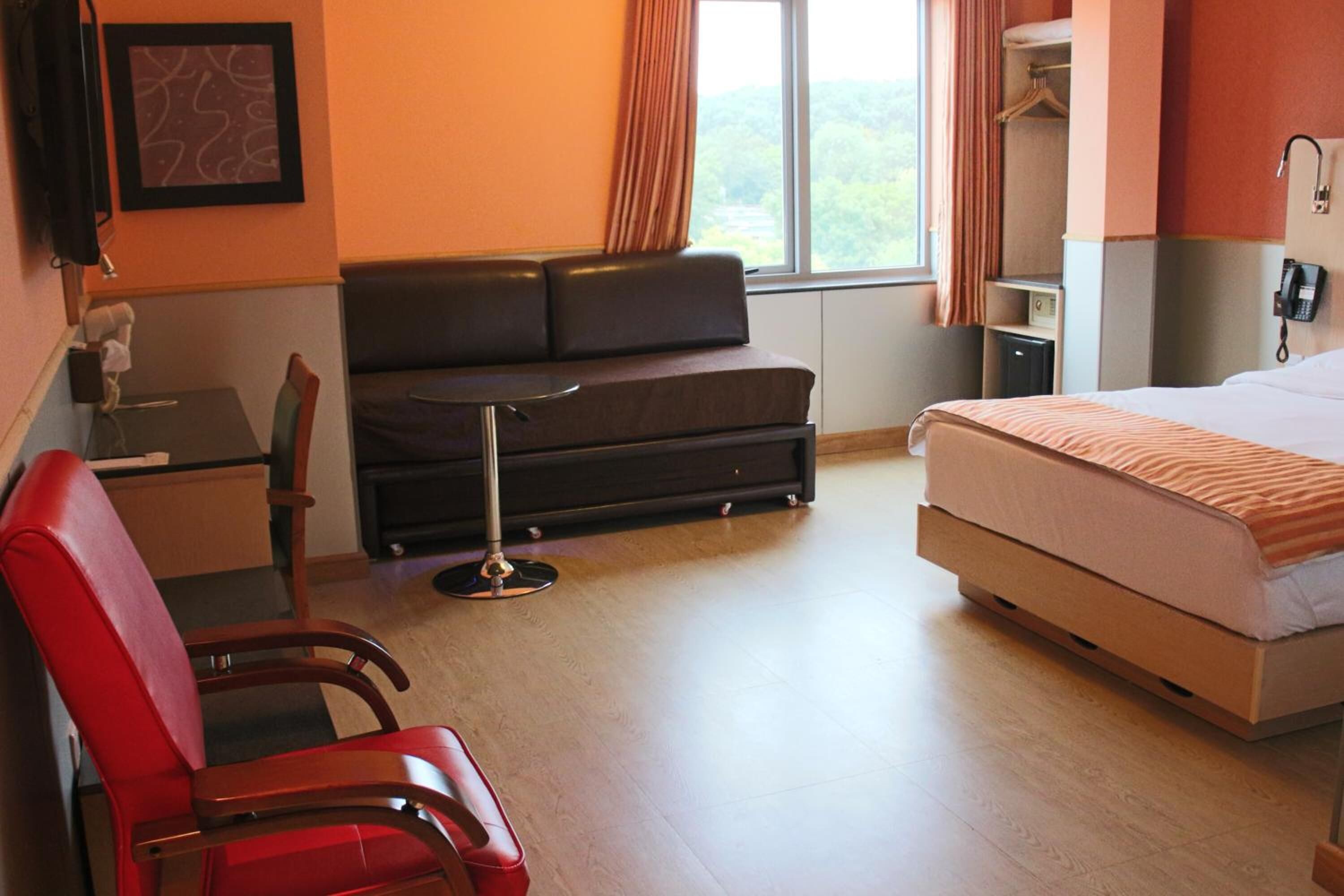 A hotel room with a bed, a sofa, a red chair, a small table, a telephone, and a window with curtains.