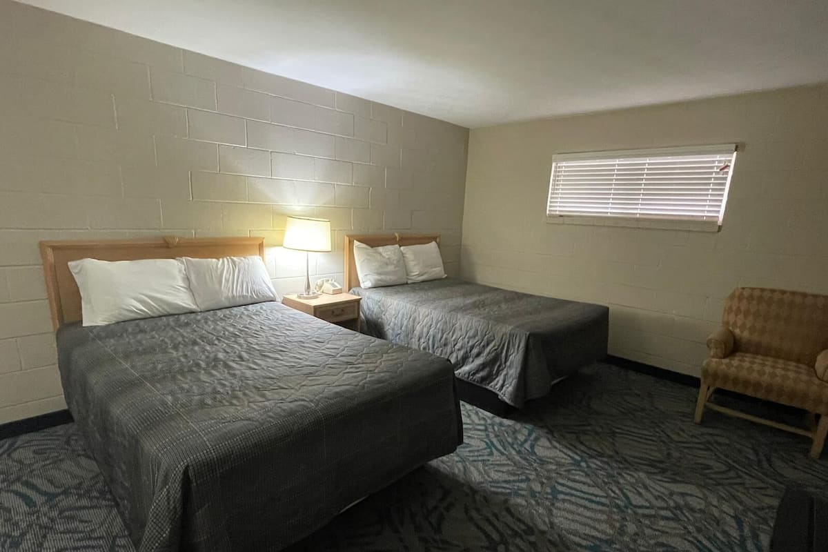 A hotel room with two beds, a nightstand with a lamp, a window with blinds, and a patterned carpet.