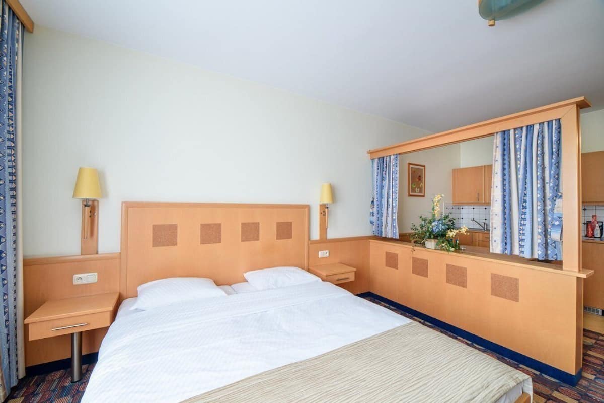 Una habitación de hotel con cama, mesitas de noche, un espejo y una kitchenette pequeña.