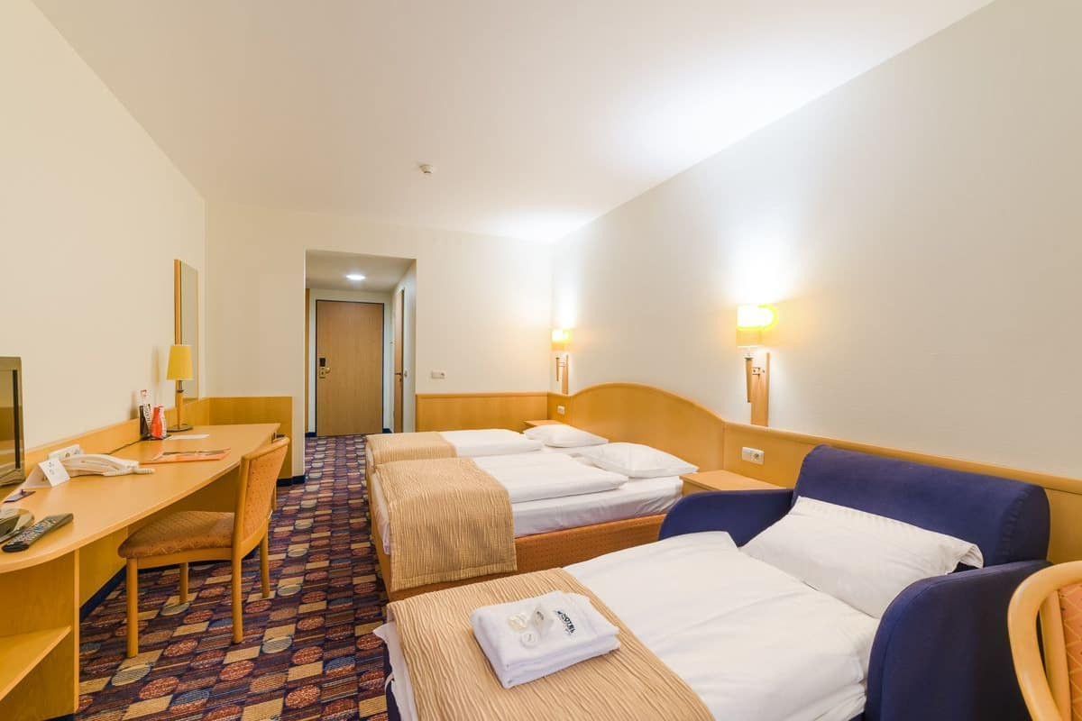 Habitación de hotel con tres camas, un escritorio y una silla. Se observan dos lámparas fijadas a la pared y una puerta al fondo.