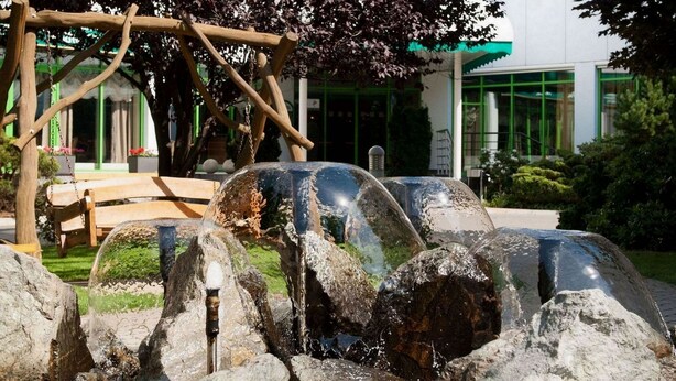 A water feature with rocks and a wooden structure in a landscaped area with a building in the background. - Hotel Partner (Warsaw)