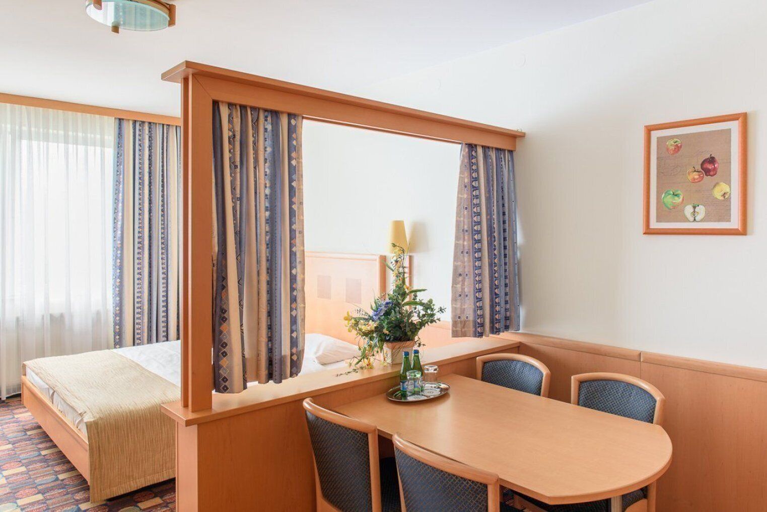 Habitación de hotel con cama, escritorio con sillas, espejo, ventana con cortinas e imagen colgada en la pared.