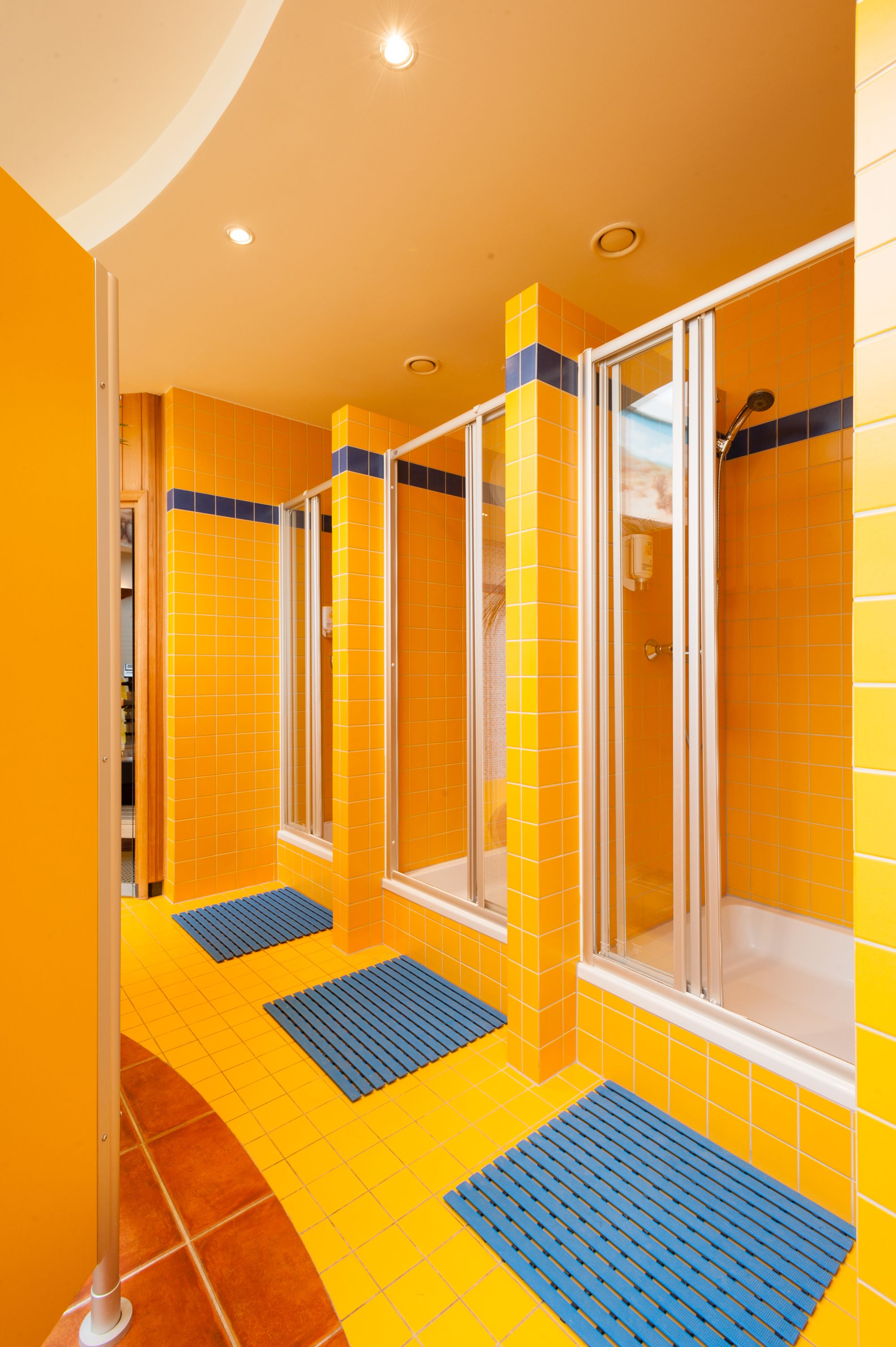 Un baño con paredes de azulejos amarillos, cabina de ducha de vidrio y una alfombra azul en el suelo.