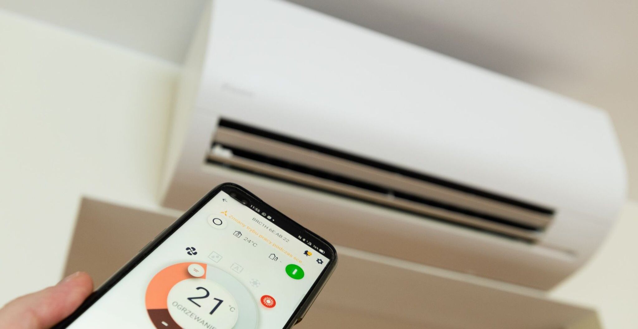 Ein Smartphone zeigt 21 Grad Celsius an, im Hintergrund ist eine Klimaanlage zu sehen.