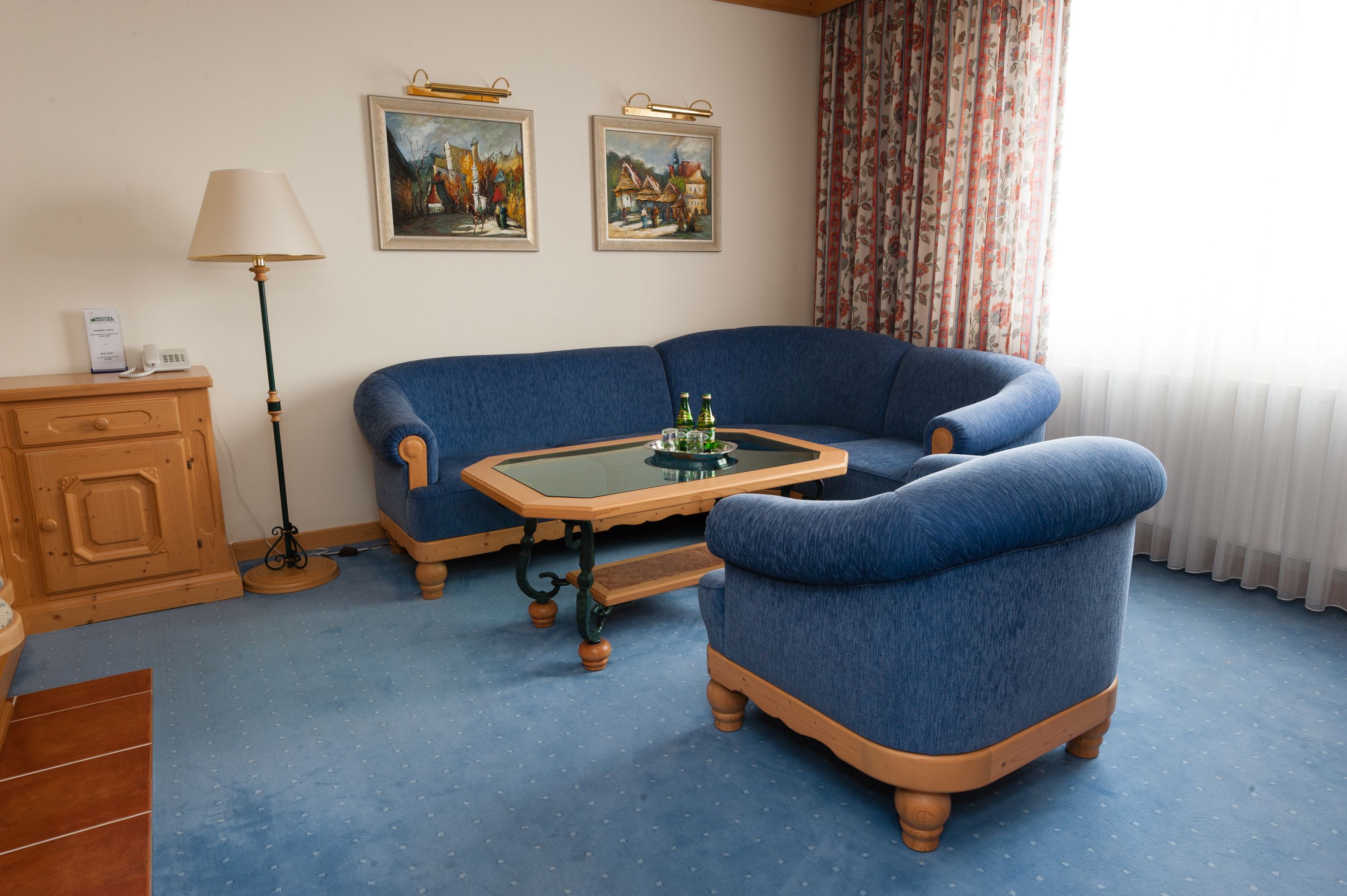 Una habitación con un sofá azul, una mesita de noche de madera, una mesa de centro de vidrio y dos cuadros en la pared.