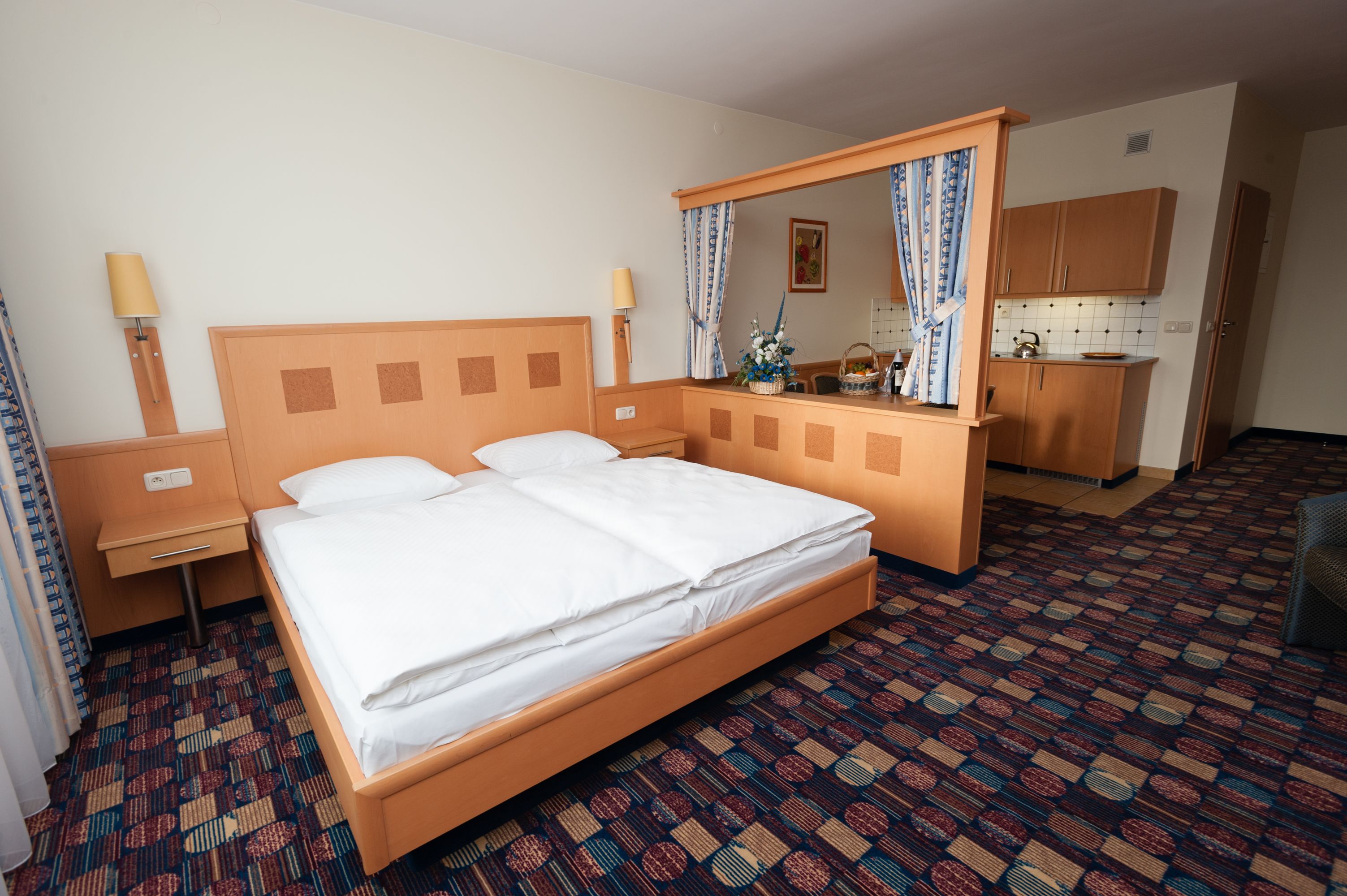 Habitación de hotel con una cama individual, una mesita de noche, un espejo, un escritorio pequeño y una zona de kitchenette.