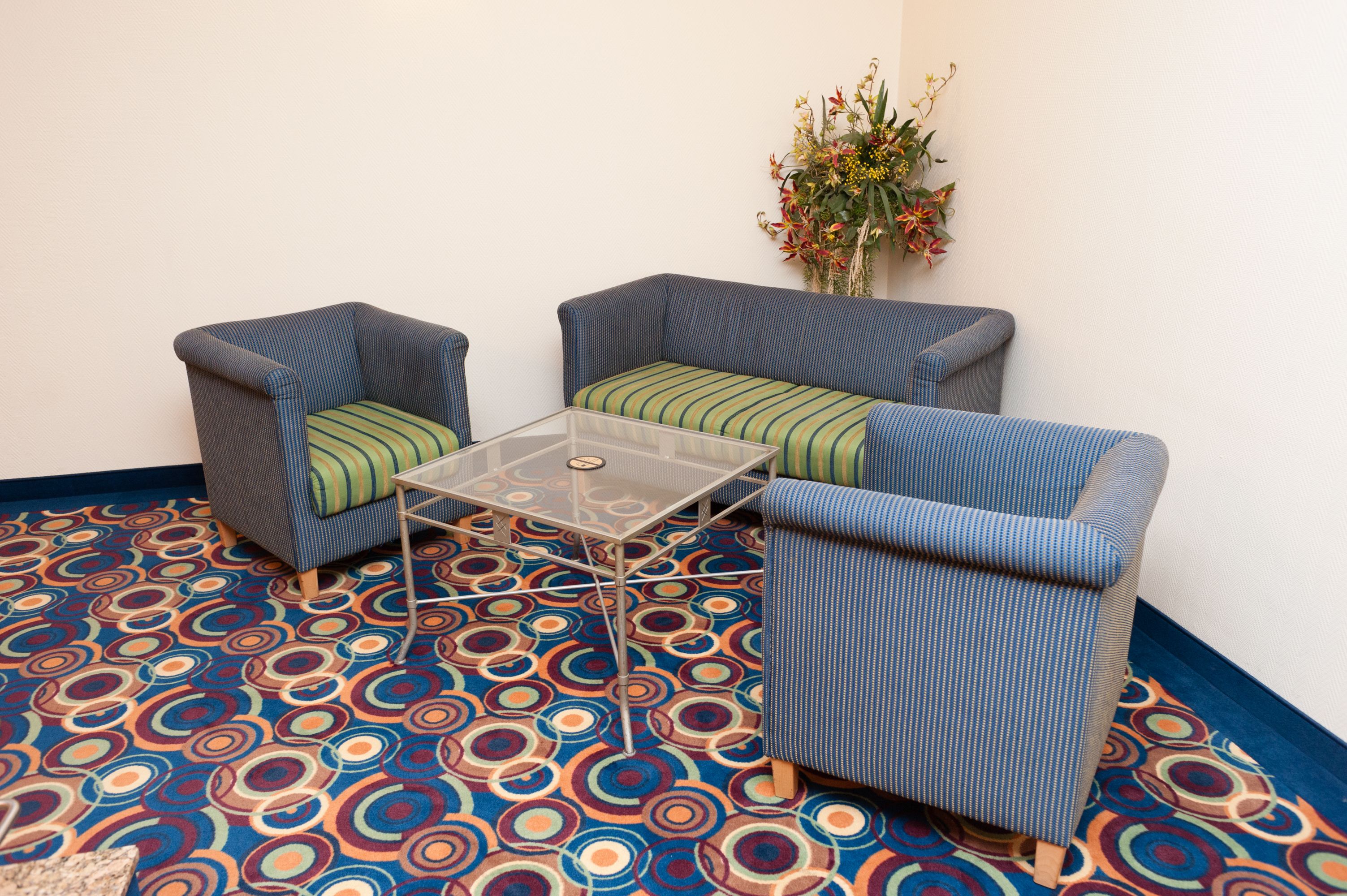Une pièce comprenant deux fauteuils et une table basse en verre, un tapis à motifs et une composition florale accrochée au mur.