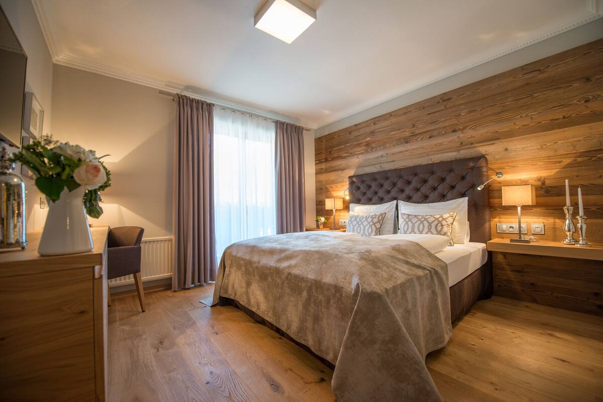 Een slaapkamer met een groot bed, een houten hoofdbord, bedlampen, een nachtkastje, een stoel en een raam met gordijnen.
