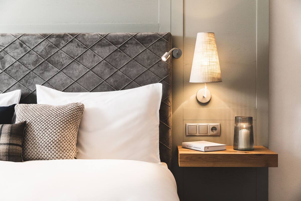 Een net opgemaakt bed met witte en gemêleerde kussens, een gestructureerd hoofdbord, een nachtkastje met een lamp en een boek.