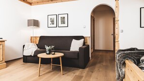 Een gezellige woonkamer met een donkere bank, een klein rond tafeltje met een vaas, houten balken in het plafond en een gang naar een andere kamer.