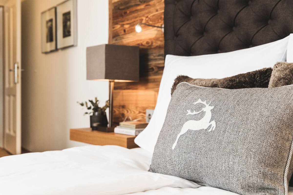 Een net opgemaakt bed met witte lakens, een donker gewatteerd hoofdbord en een decoratief kussen met een hertenprint.