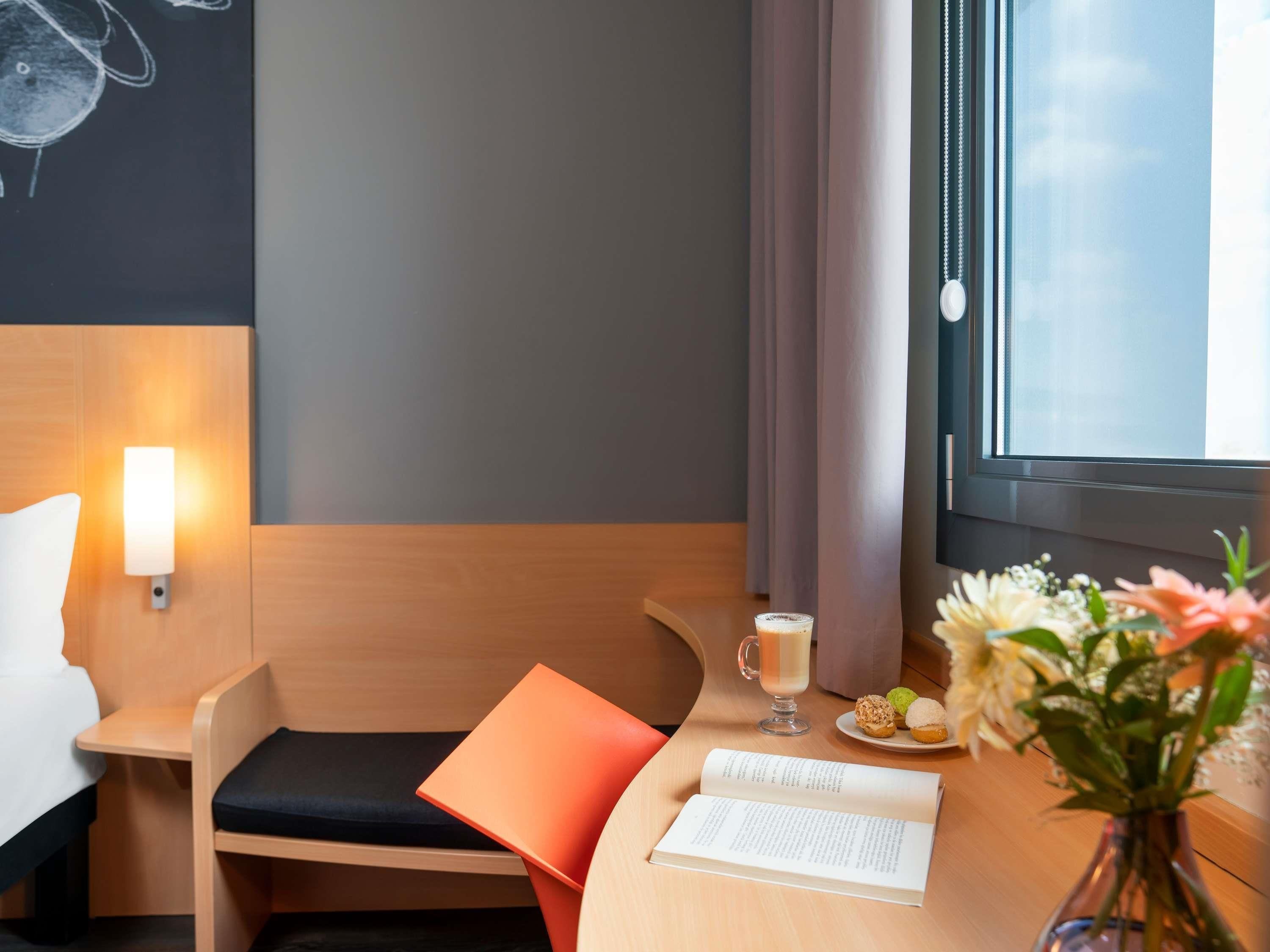 Une chambre d’hôtel avec un lit, un bureau avec un ordinateur portable, une tasse de café, une assiette de viennoiseries et un vase rempli de fleurs.