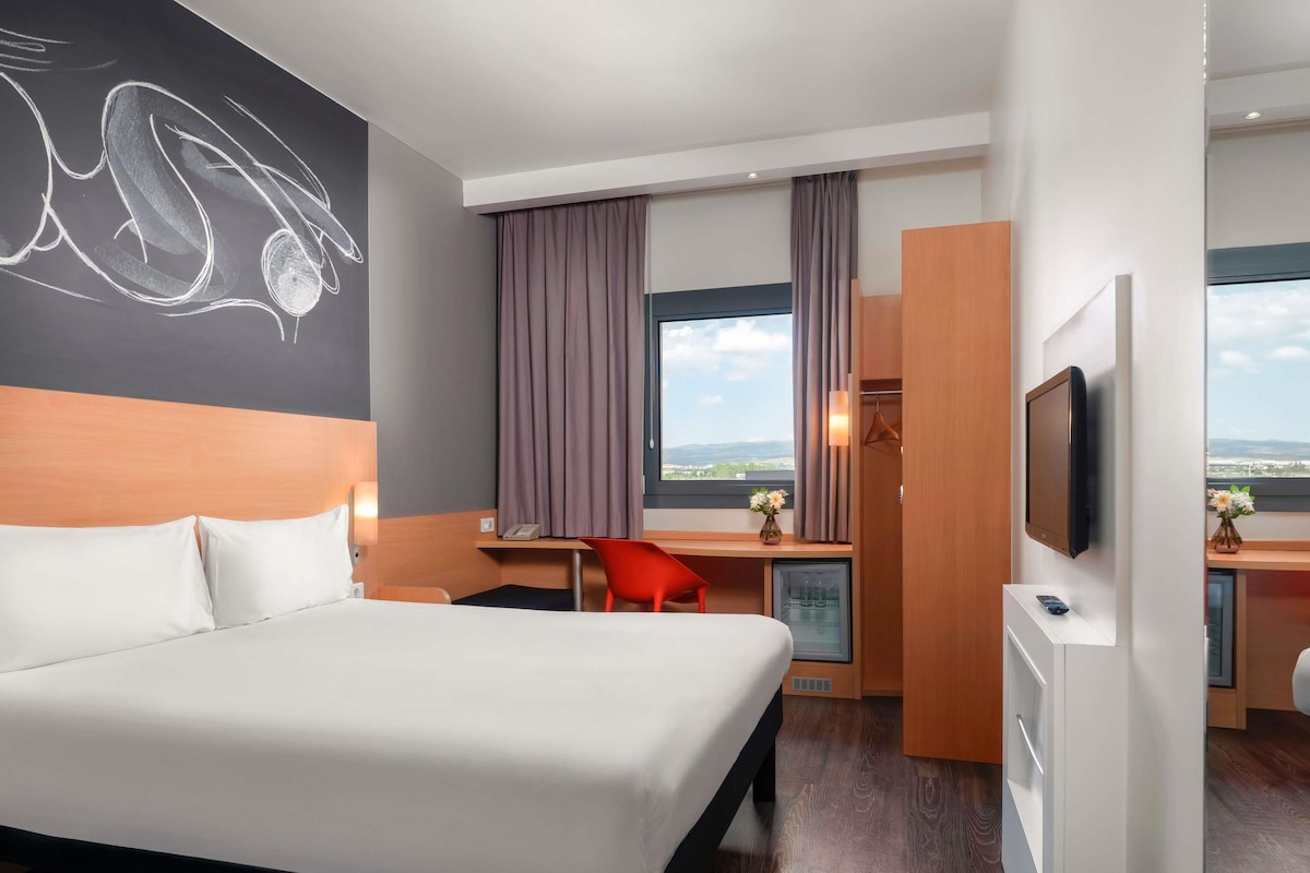 Habitación de hotel con cama, escritorio, silla, televisión y vista a la ciudad.