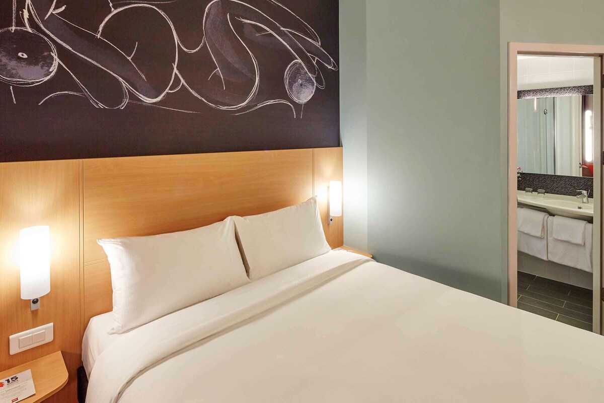 Een hotelkamer met een groot bed, twee bedlampen, een houten hoofdbord en een badkamer die door een open deur zichtbaar is.