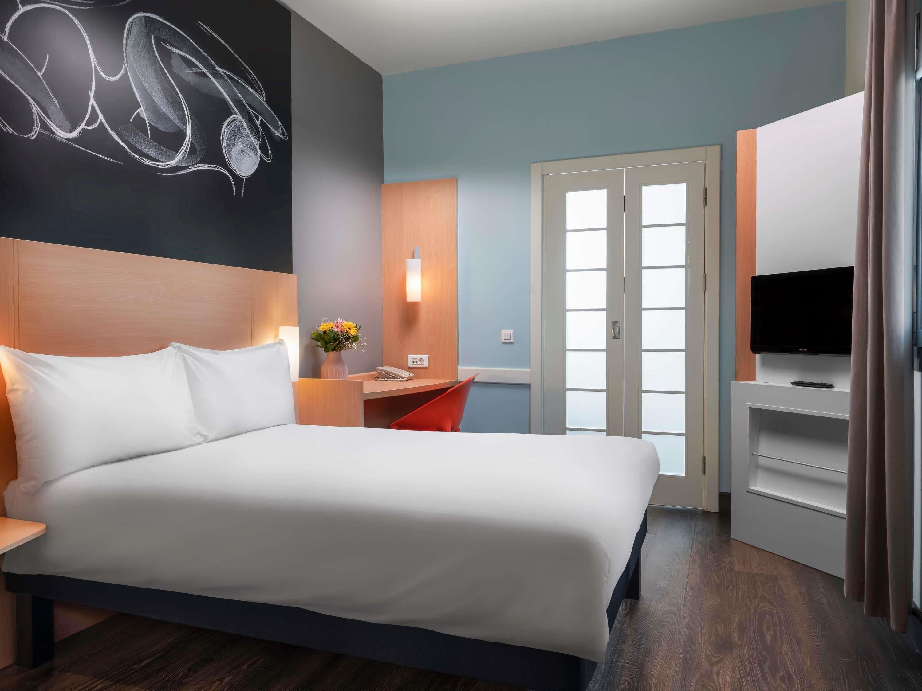 A hotel room with a large bed, a desk with a chair, a flat-screen TV, and a door leading to a balcony.