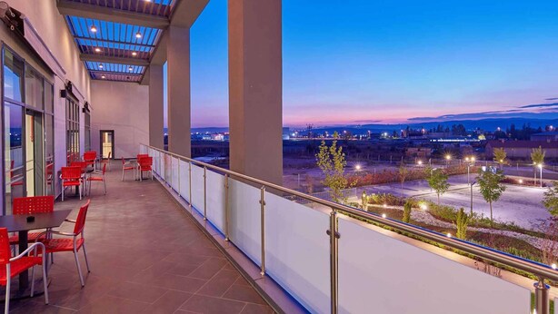 Un balcon avec des chaises et des tables rouges, un plafond en verre et une vue sur le paysage urbain au crépuscule.