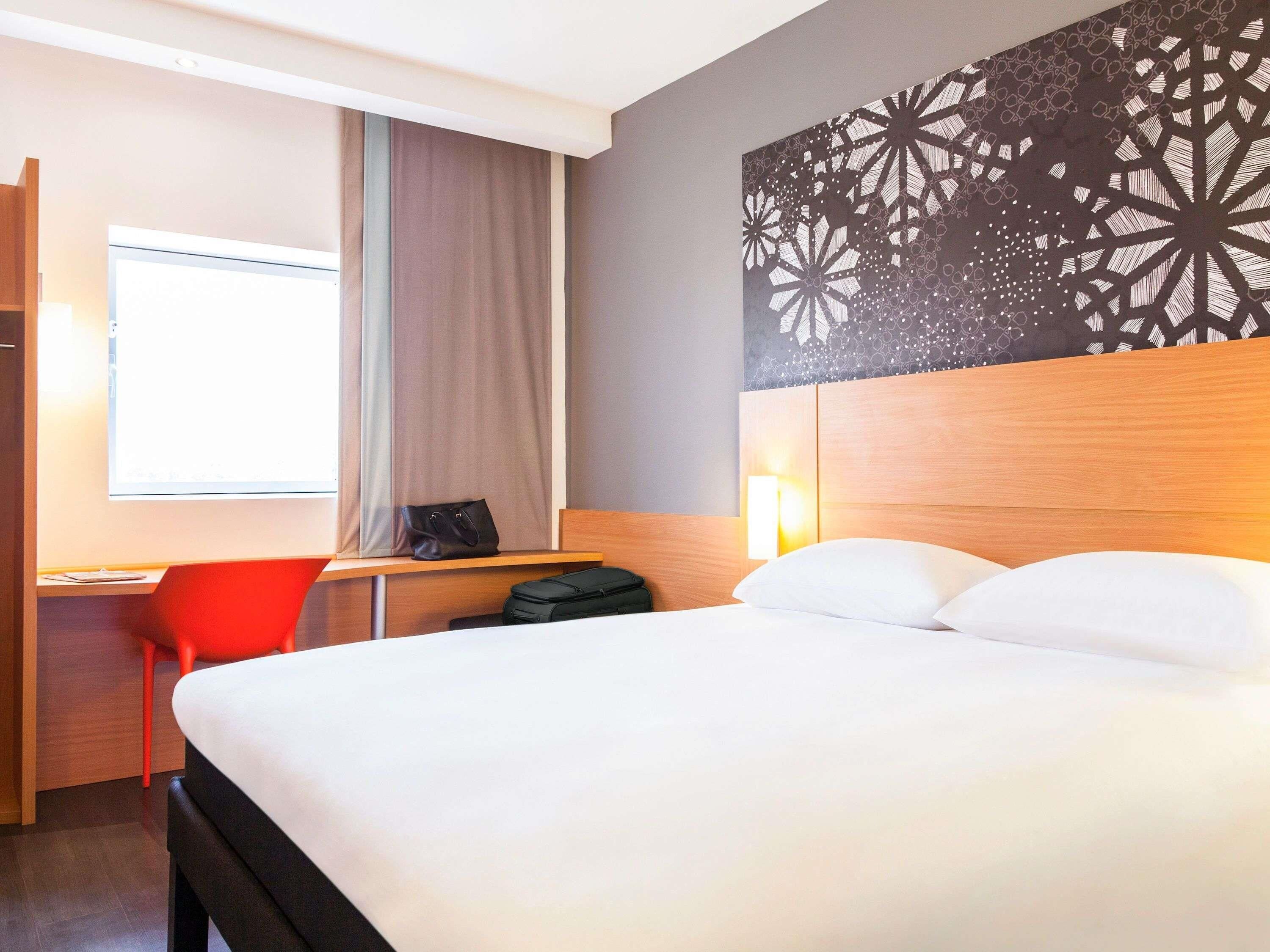 A hotel room with a bed, a desk with a chair, a window with curtains, and a wall with a geometric pattern.