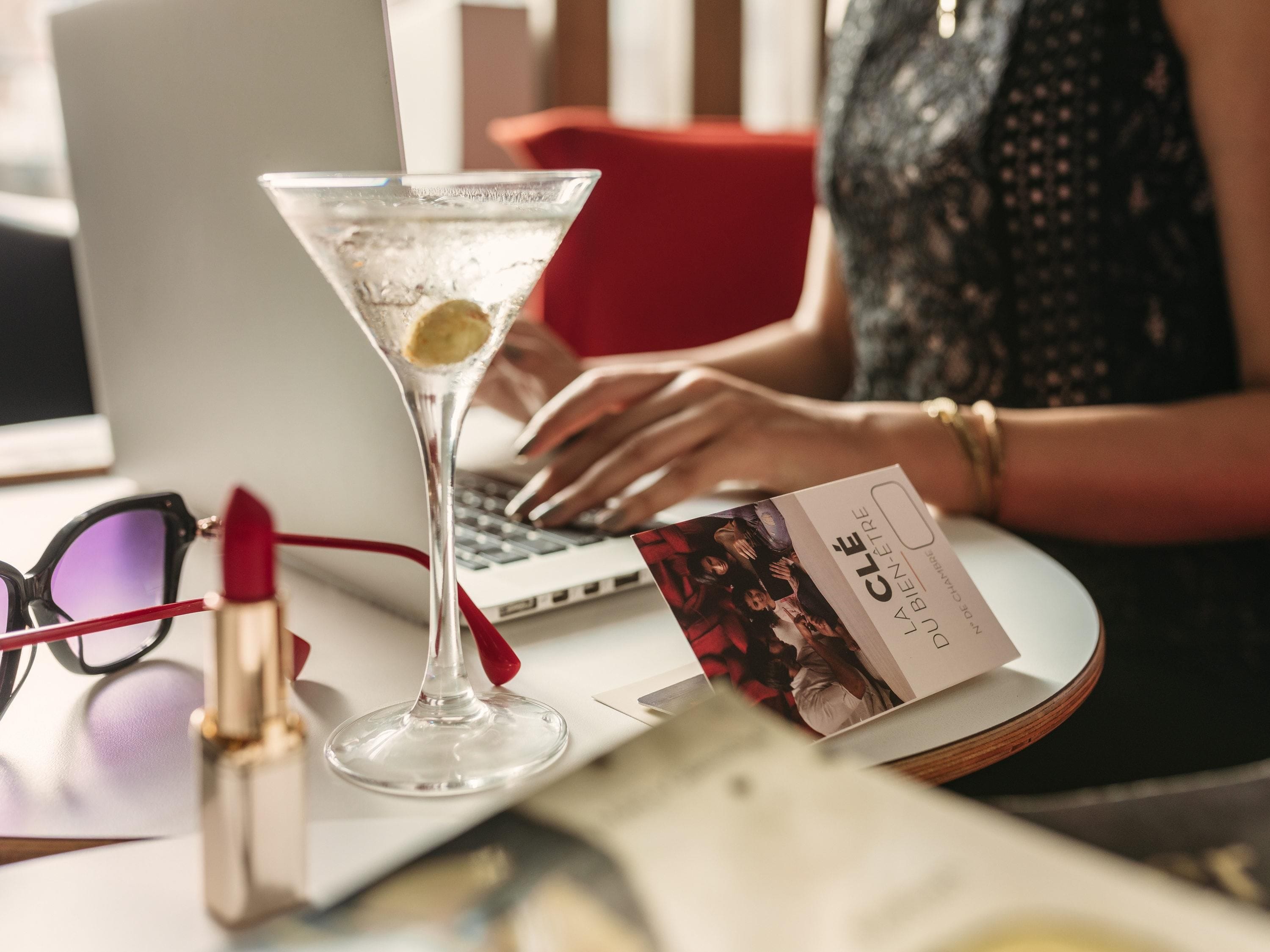 Ein Laptop, ein Martini-Glas mit Zitronenscheibe, ein Lippenstift und eine Visitenkarte auf einem Schreibtisch.