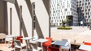 Un espace extérieur aménagé avec des tables et des chaises, un bâtiment moderne en arrière-plan et un ciel dégagé.