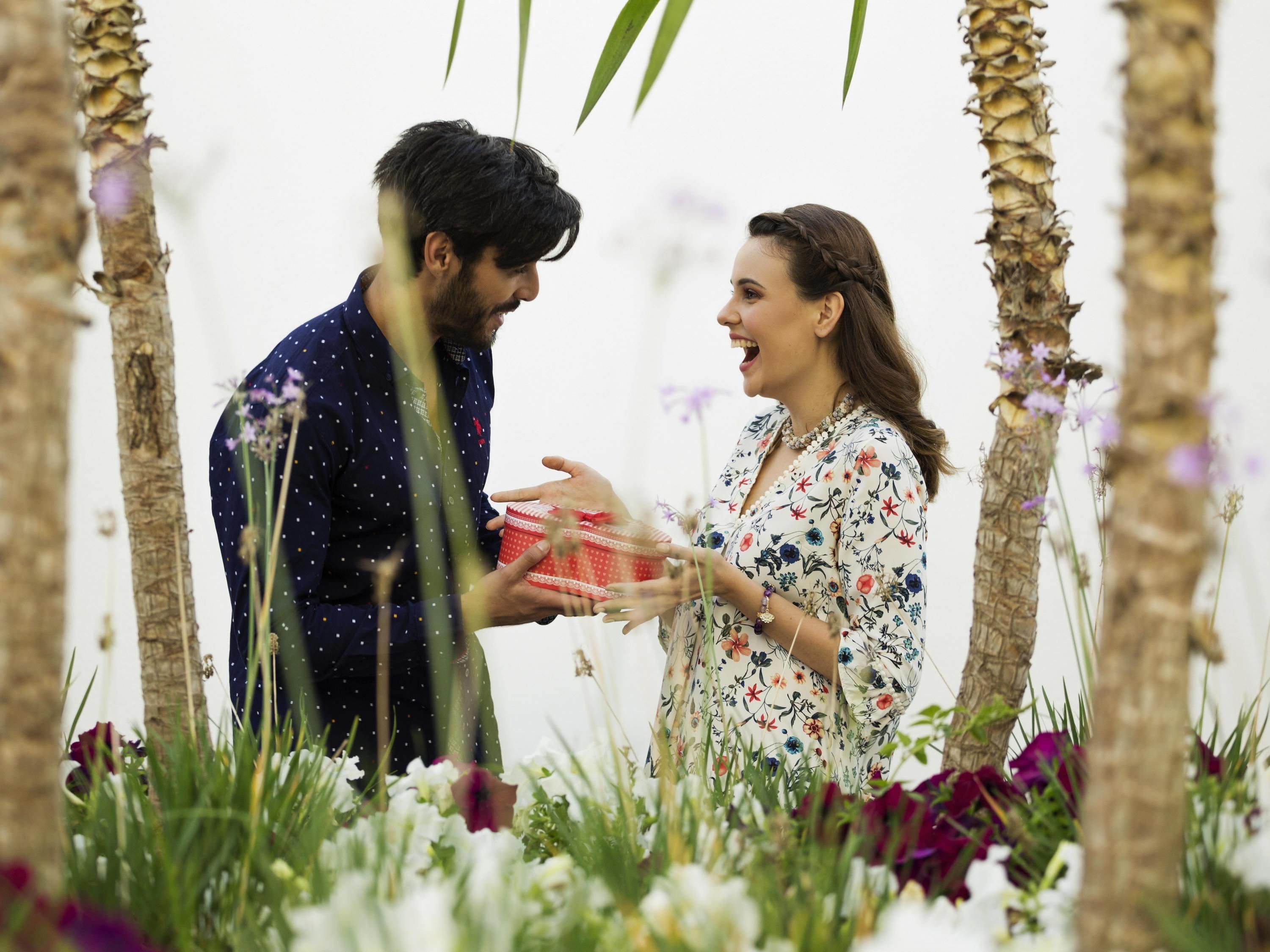 Een stel dat elkaar een cadeau geeft, in een tuin met palmbomen en bloemen.