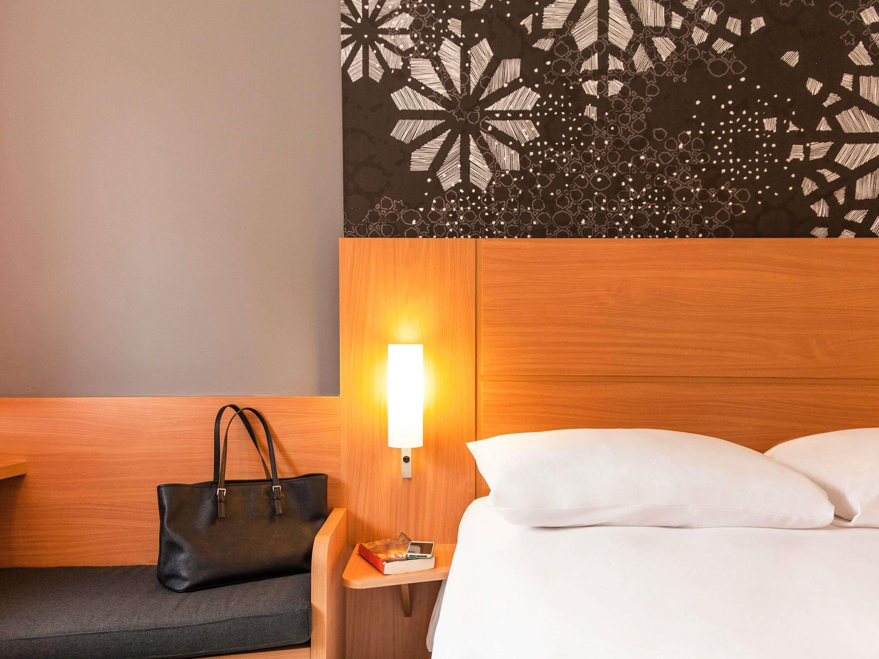 A hotel room with a wooden headboard, a bedside lamp, a black leather bag, a book, and a white bed.