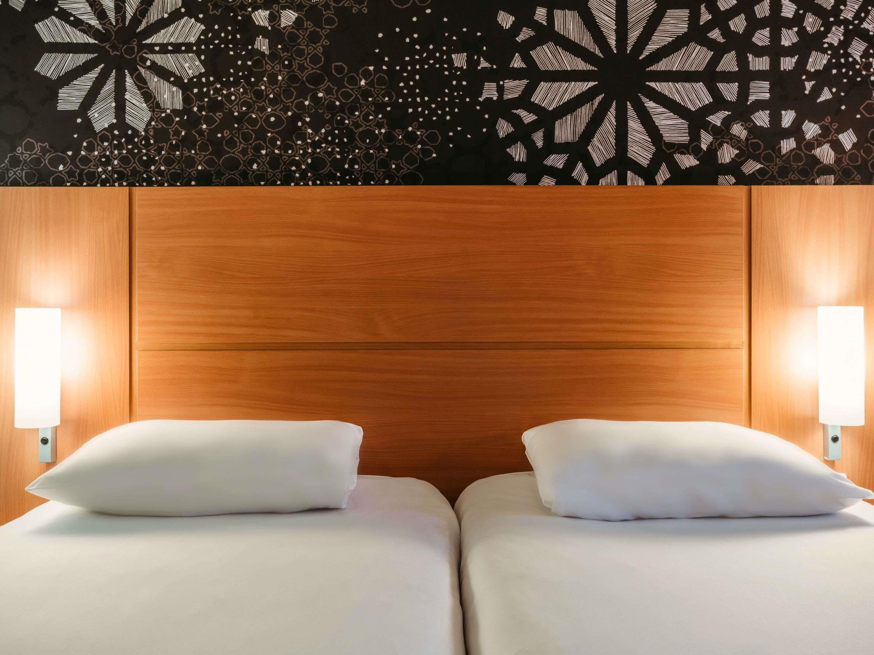 A hotel room with a wooden headboard, two beds with white linens, and wall-mounted lamps.