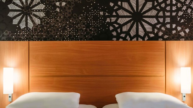 Une chambre d’hôtel avec une tête de lit en bois, deux lits avec du linge de lit blanc et des lampes fixées au mur.