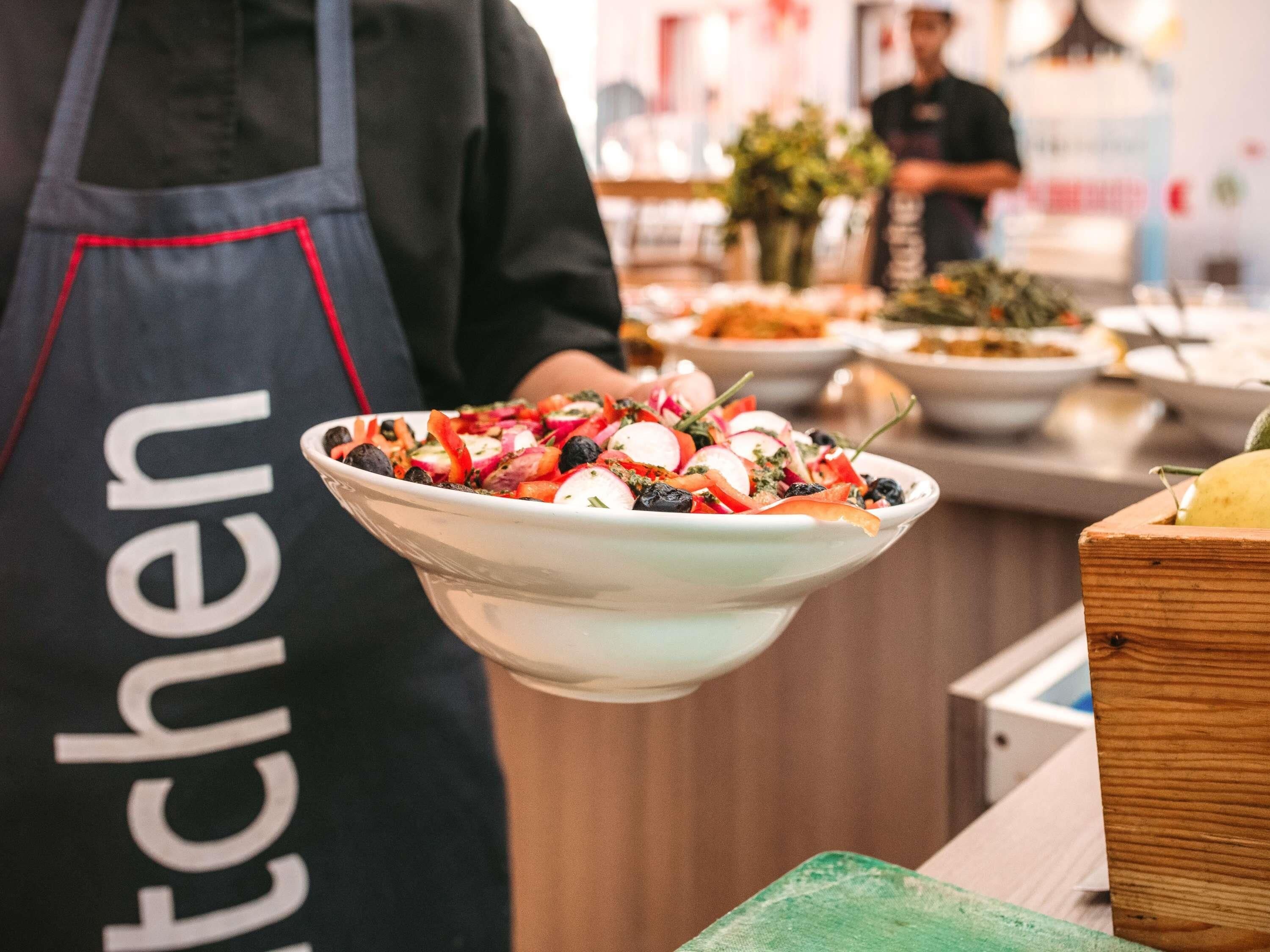 Una persona con delantal negro que tiene la palabra 'cocina' escrita, sosteniendo un tazón de ensalada.