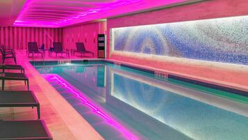 Una piscina interior moderna con iluminación rosa, agua cristalina y sillas de descanso a un lado.