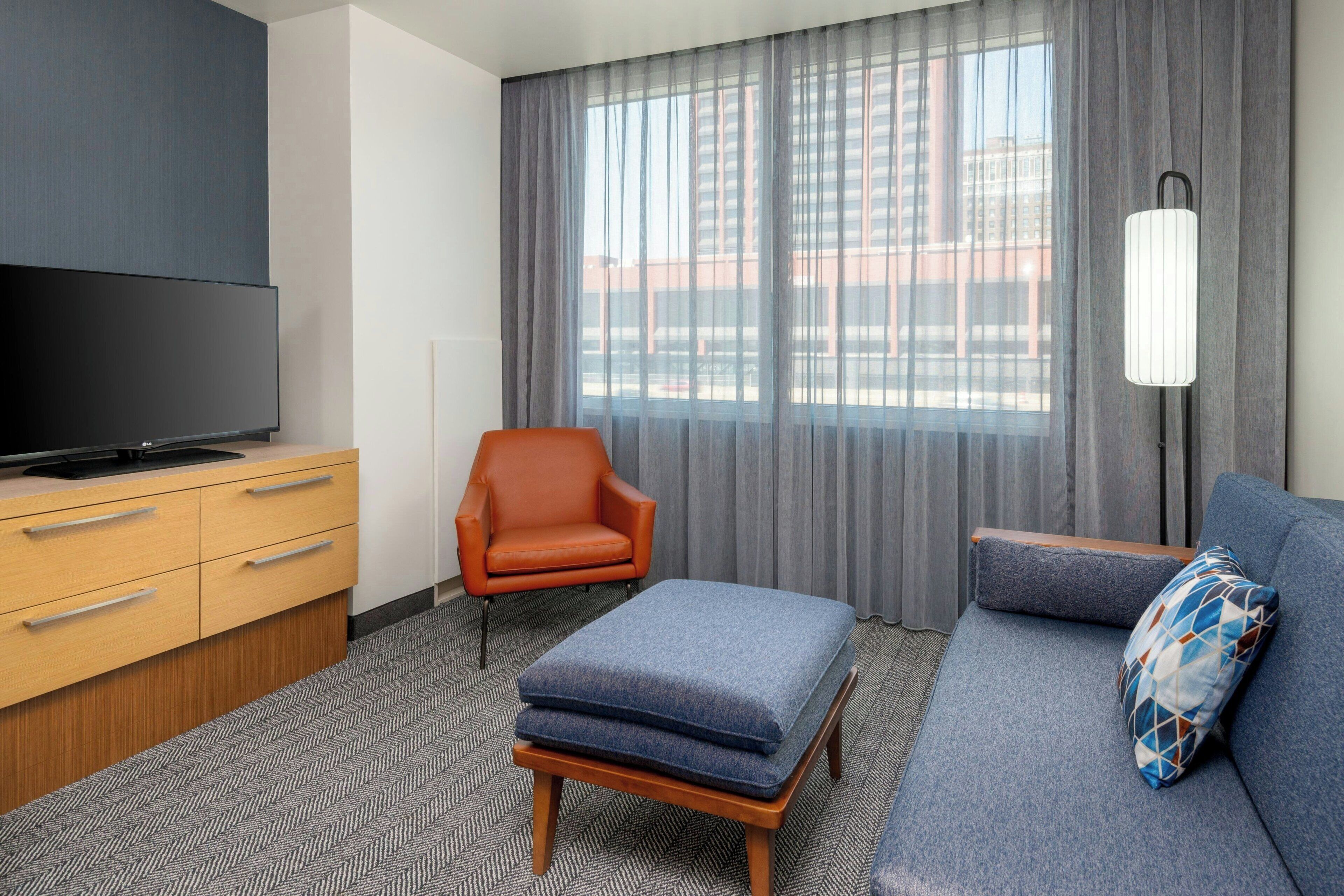 A modern hotel room with a flat-screen TV, a wooden cabinet, a blue sofa, an orange armchair, and a large window with curtains.
