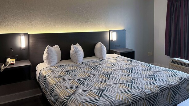 Una habitación de hotel con una cama grande, dos mesitas de noche, un buró, una lámpara de pared y una ventana con cortinas.