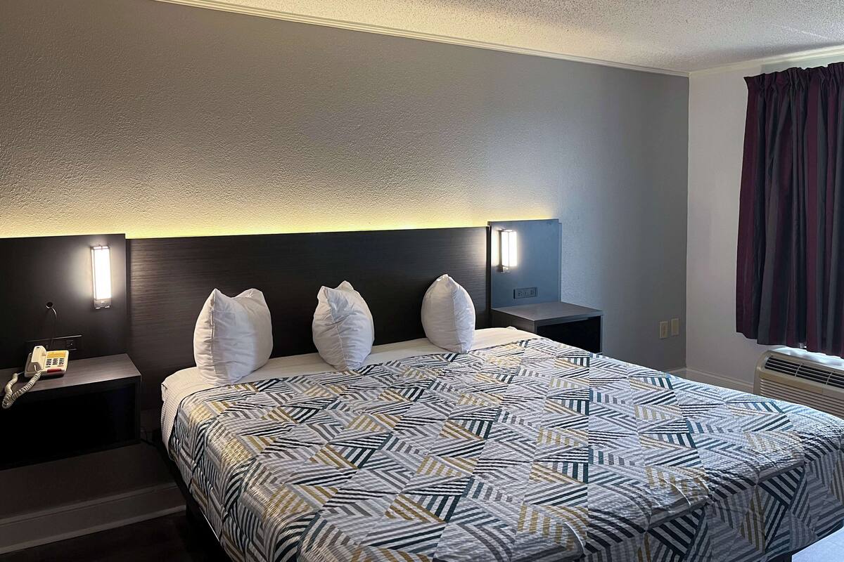 A hotel room with a large bed, two bedside tables, a nightstand, a wall-mounted lamp, and a window with curtains.