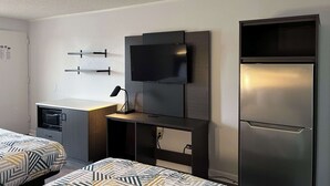 Una habitación de hotel moderna con dos camas, un televisor de pantalla plana montado en un panel de madera, un microondas integrado y un refrigerador de acero inoxidable.