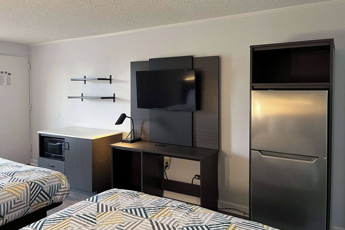 A modern hotel room with two beds, a flat-screen TV mounted on a wooden panel, a built-in microwave, and a stainless steel refrigerator.