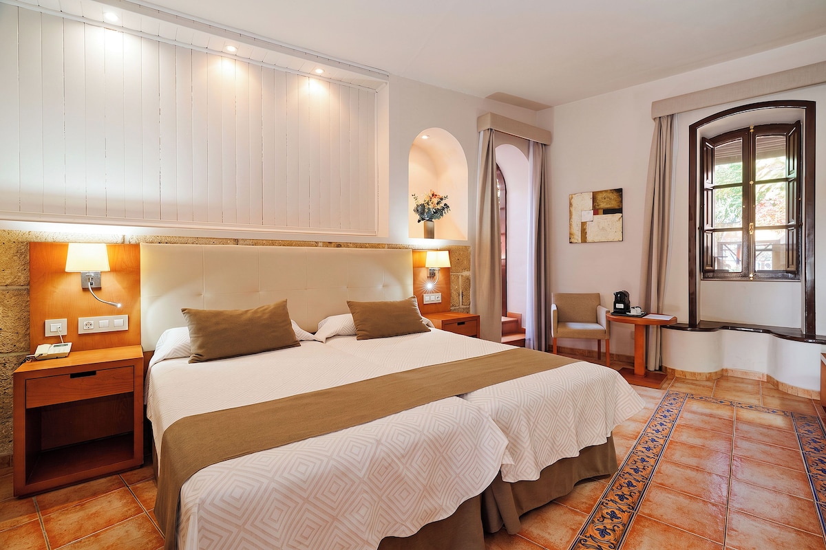 A hotel room with a large bed, bedside tables, a desk, a chair, a window with curtains, and a decorative wall piece.