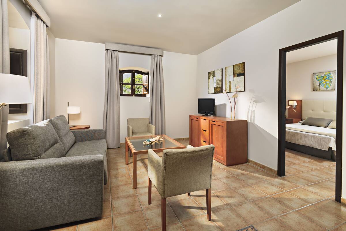Une chambre d’hôtel moderne avec un canapé, un fauteuil et une petite table. On y trouve également une télévision, une armoire et un lit avec une tête de lit.